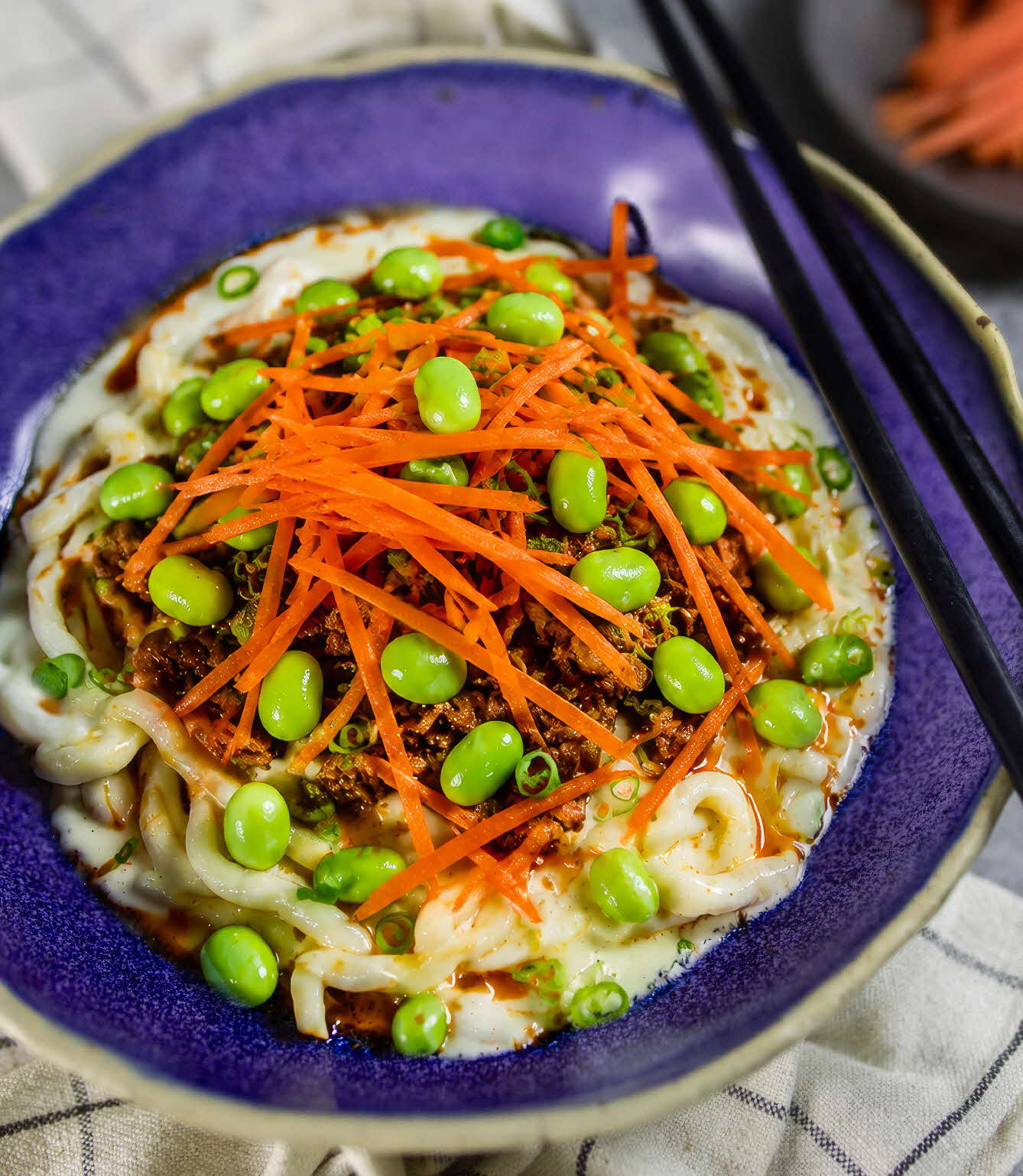 A bowl of creamy tofu udon topped with minced chicken, edamame, carrots and spring onions.