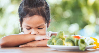 A grumpy young kid with her arms folded on the table, refusing to eat her vegetables.