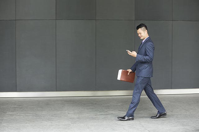 businessman casually walking looking at his phone while holding a folder in his other hand