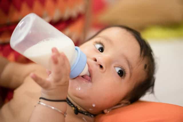 baby drinking either breast milk or formula milk from a baby bottle, because milk should still be the main source of nutrition for a baby when introducing solid foods.>