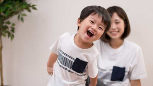 Child smiling while playing as a mother sits nearby in a home setting.