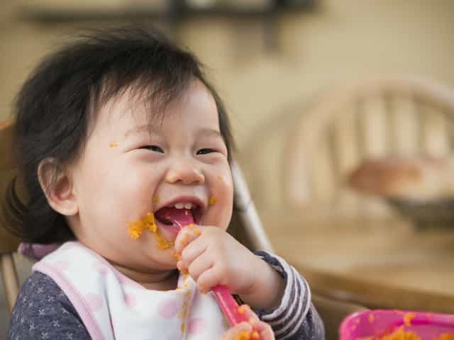 Baby holding a baby spoon in her mouth and smiling. These are signs baby is ready for solid food.>