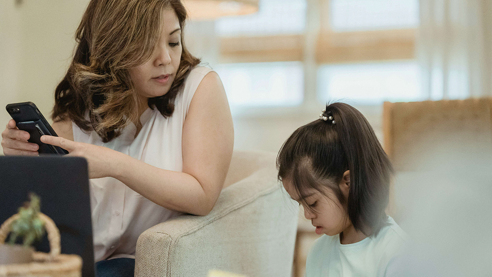 Mother using a smartphone while a child sits nearby at home.