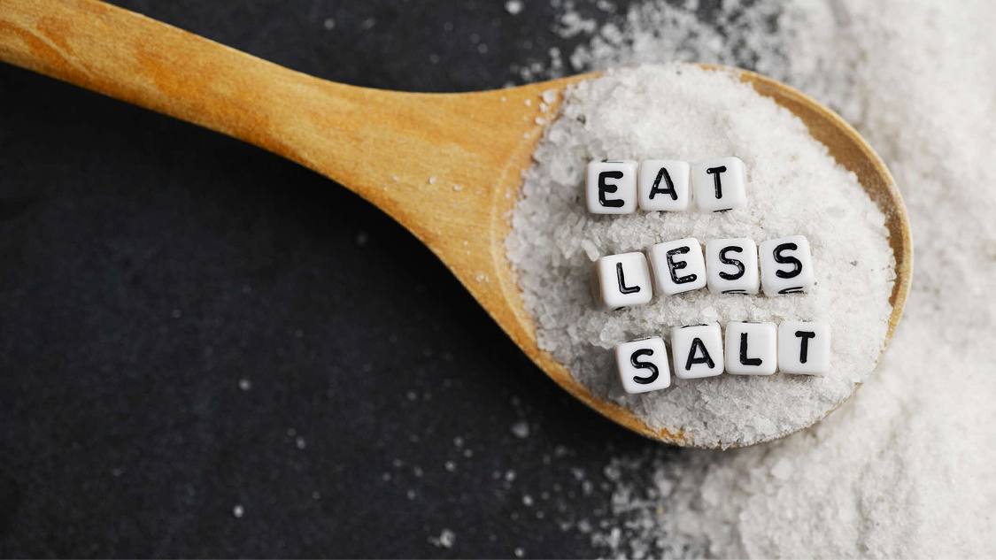 Wooden spoon filled with salt and letter tiles spelling “Eat Less Salt”.