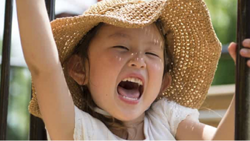 Child playing outdoors, smiling and raising arms while wearing a sun hat.