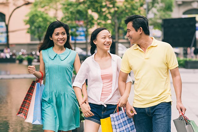 asian daughter smiling and linking arms with her father and mother while out shopping