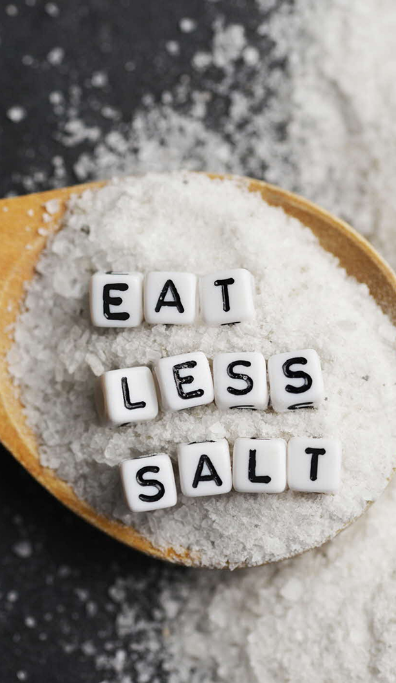 Wooden spoon filled with salt and letter tiles spelling “Eat Less Salt”.