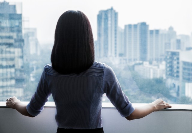 asian woman in office attire peering out a window